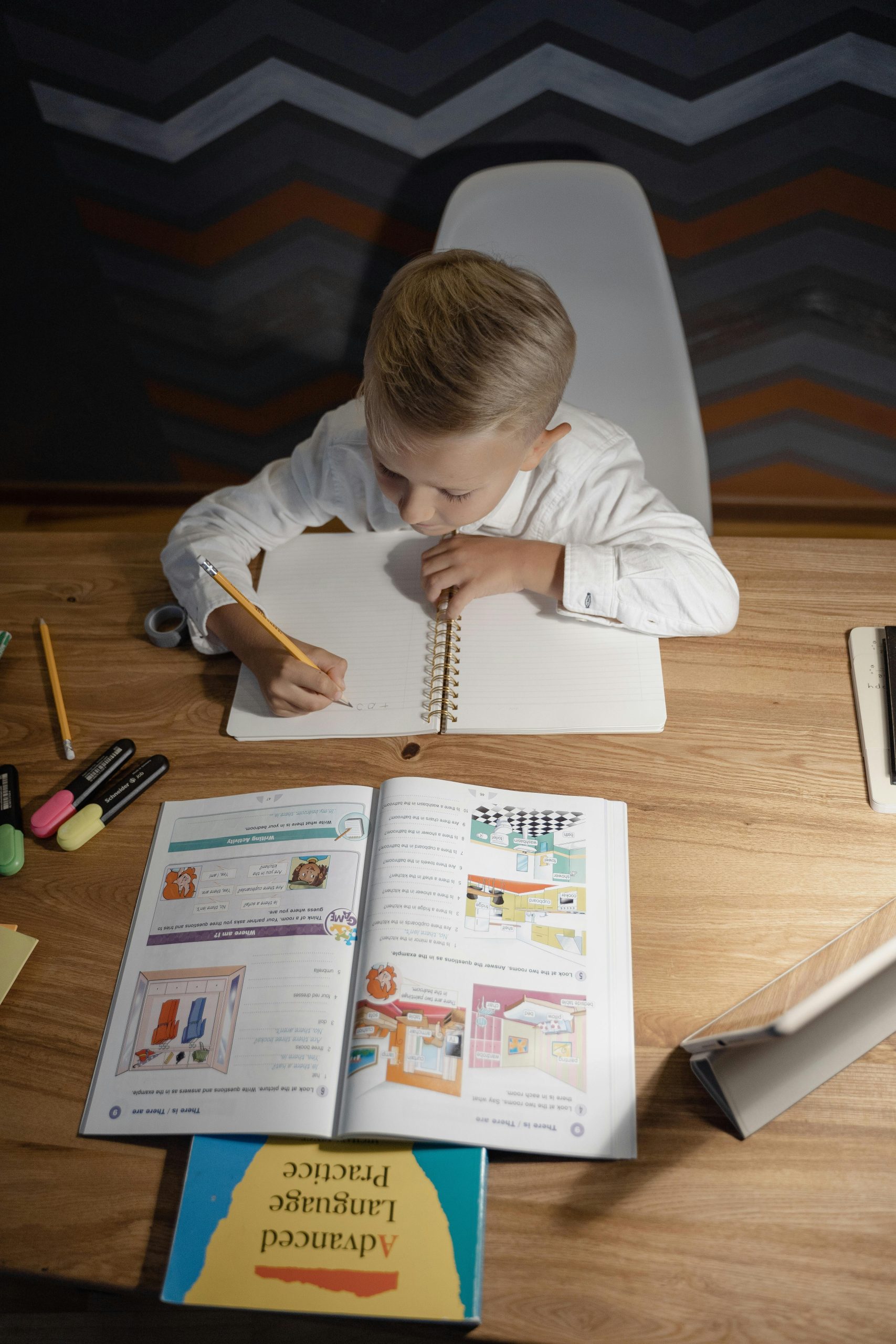 A child focused on homework, writing in a notebook at a wooden desk, surrounded by study materials.